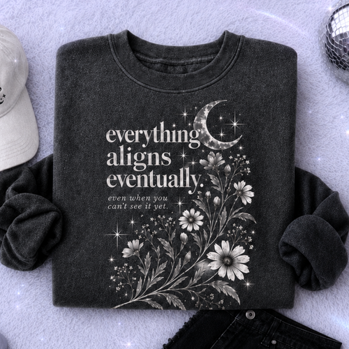 A styled flat-lay featuring a dark gray graphic t-shirt with white text and floral/crescent moon artwork. The shirt is surrounded by coordinating accessories including a white baseball cap, black distressed shorts, a silver star-shaped accessory, a small silver phone, and a lavender notebook on a sparkly lavender background.